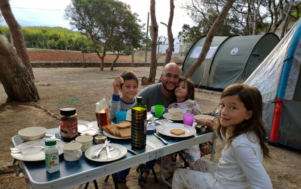 Foto capturada por Tere Watson de su marido y sus tres hijos mayores desayunando juntos en una mesa portátil en un camping. La imagen muestra un momento íntimo y alegre de la familia compartiendo tiempo juntos al aire libre, reflejando la simplicidad y la felicidad de las experiencias de viajes en familia.