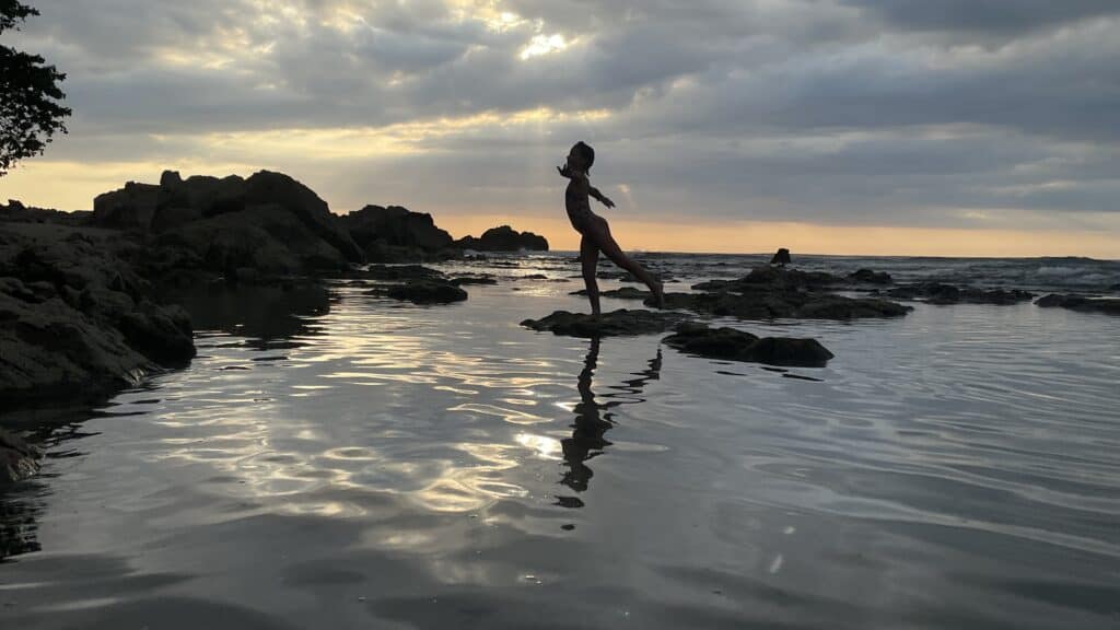 Vicky, hija de Tere Watson, haciendo equilibrio sobre una roca en la playa al atardecer durante un viaje en familia en Costa Rica. La imagen captura un hermoso momento al atardecer frente al mar, simbolizando la paz y la libertad que disfrutan en sus viajes familiares, rodeados de un paisaje natural impresionante.