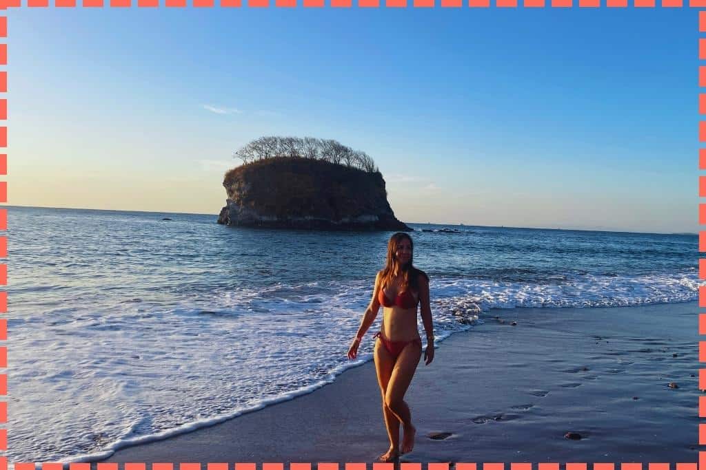 Mujer caminando en bikini por Bahía de los Piratas, disfrutando del atardecer con el islote al fondo en las playas de Costa Rica.