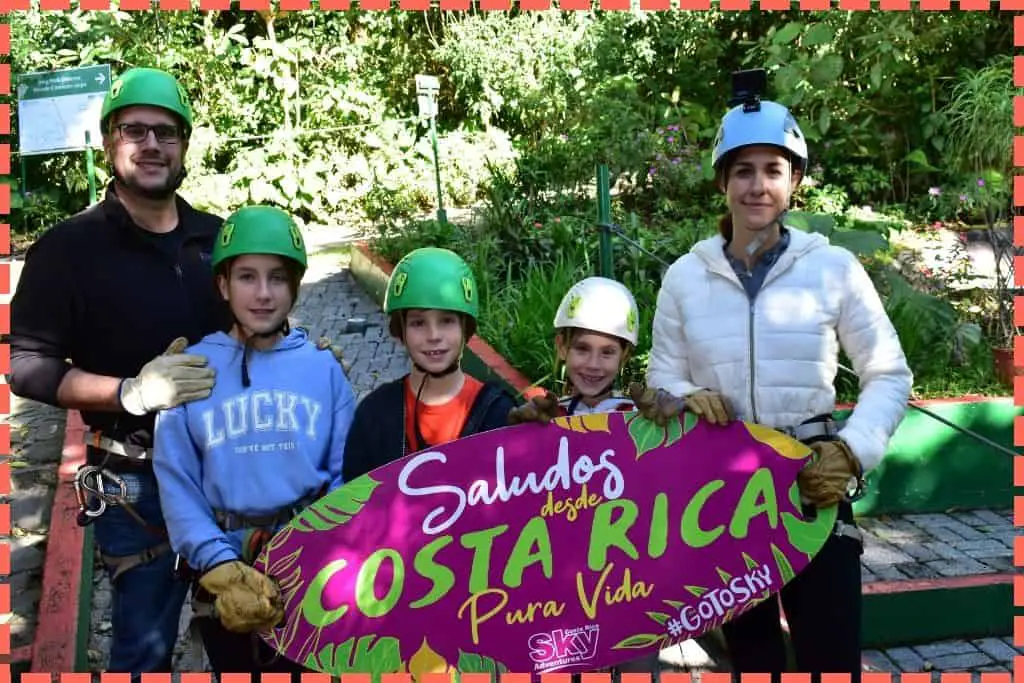 Familia Watson con equipo de canopy de Sky Adventures en Monteverde. Sosteniendo un cartel que dice 'Saludos desde Costa Rica, pura vida'.