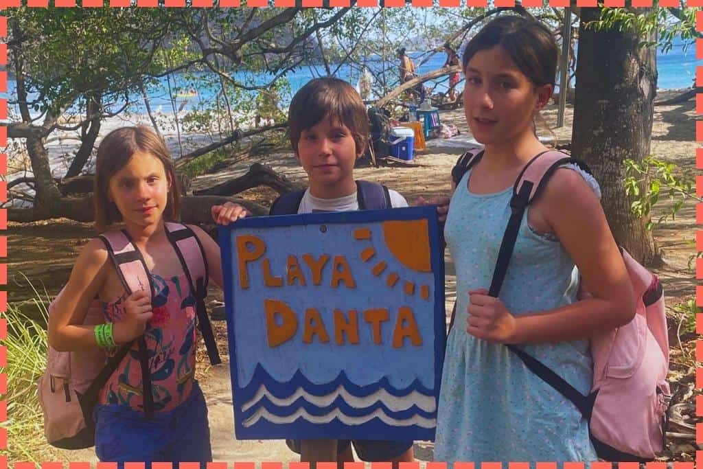 Tres niños posando con el cartel de Playa Danta, con la playa y visitantes de fondo en las playas de Costa Rica.