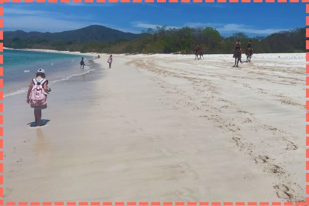 Familia Watson caminando en la orilla de arena blanca de la Playa Conchal, Guanacaste, mientras tres jinetes a caballo pasean en la misma dirección. Aguas cristalinas y montañas en el fondo. Guanacaste preciosidad en Costa Rica.