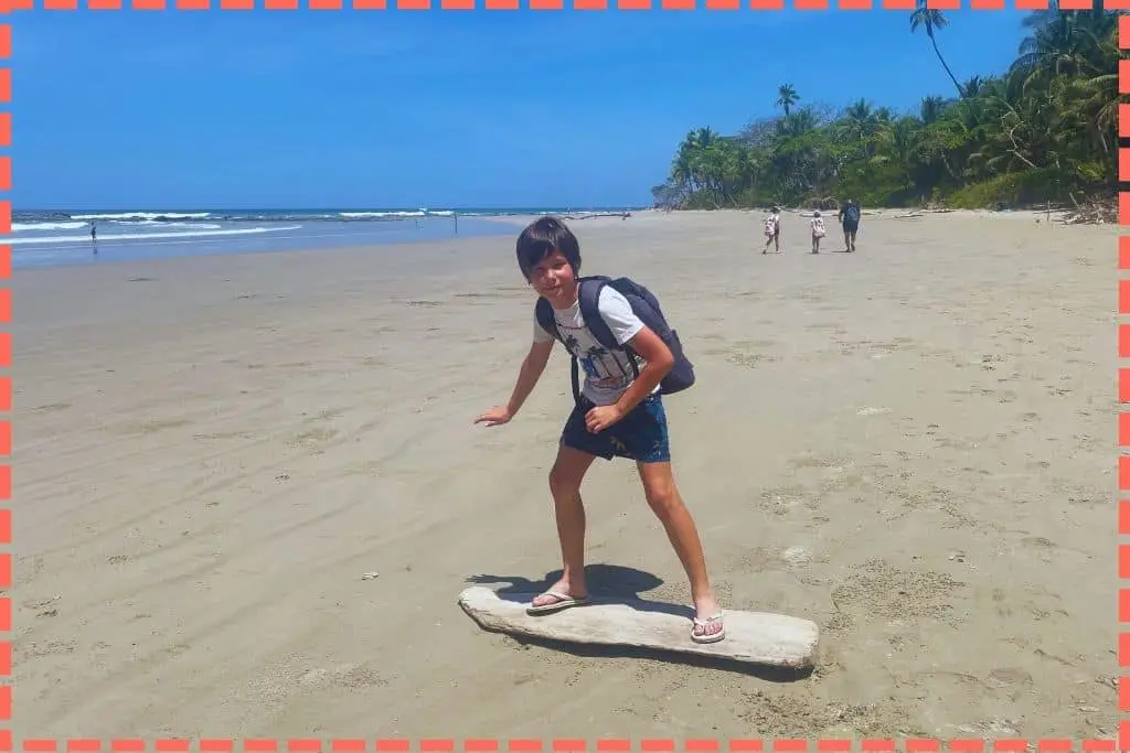 Ian sobre un tronco de madera con forma de surf en la arena, padre y dos hijas caminando de fondo, en el sur de Nicoya, Costa Rica, con selva, mar, arena y cielo azul brillante.