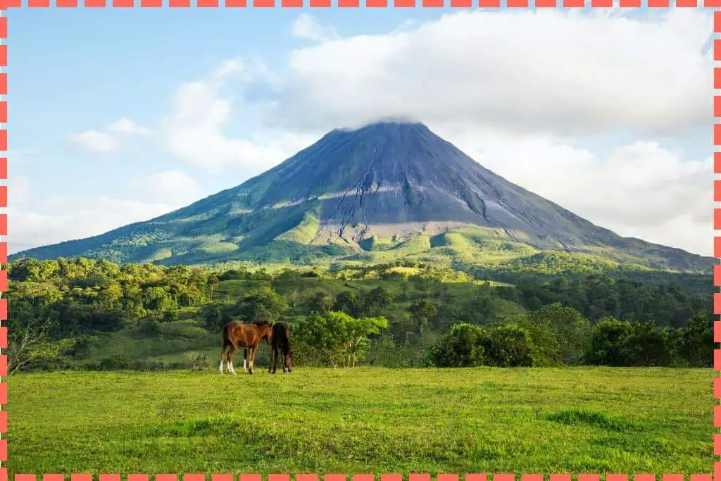 Imagen del Volcán Arenal en la Fortuna, Costa Rica, con una nube sobre él, rodeado de exuberante vegetación y dos caballos en primer plano.