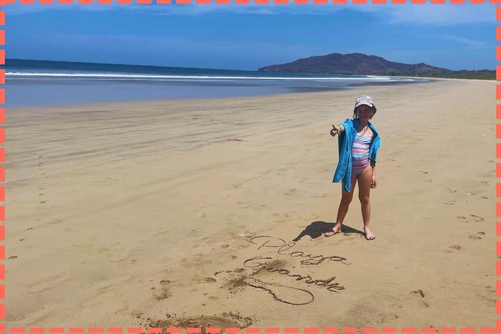 Vicky sonriente en Playa Grande, Costa Rica, con la frase 'Playa Grande' creativamente escrita en la arena a su lado.