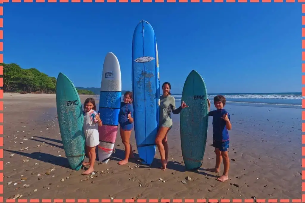 Tere Watson y sus  3 hijos con tablas de surf, gesto de surf en la playa con árboles a la izquierda y el mar a la derecha. Una de las actividades que hacer en Costa Rica.