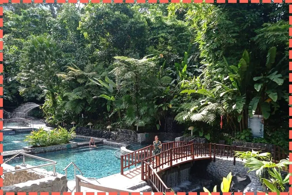 Foto en las Termas del Arenal, Costa Rica, con piscinas de diferentes temperaturas rodeadas de palmeras y selva. Una persona en un puente mira a la cámara mientras otra se relaja en segundo plano. 