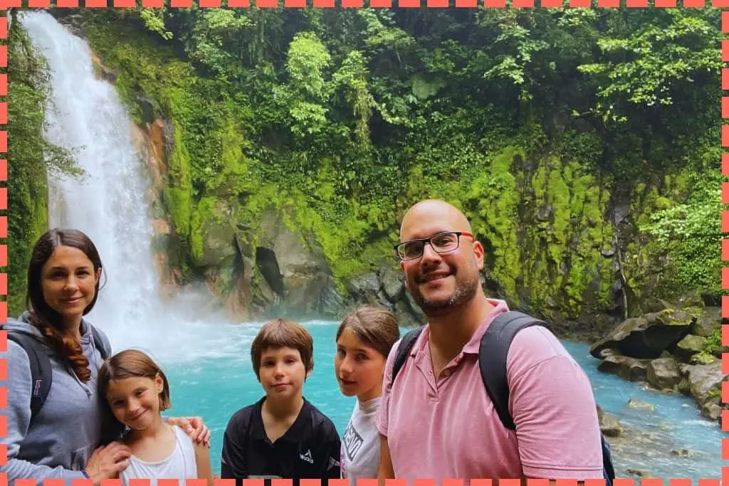 Familia de 5 personas sonríe a la cámara, disfrutando de la espectacular cascada del río Celeste, con su característico azul intenso, rodeada de rocas y vegetación exuberante. Uno de los paisajes más hermosos para visitar en Costa Rica.