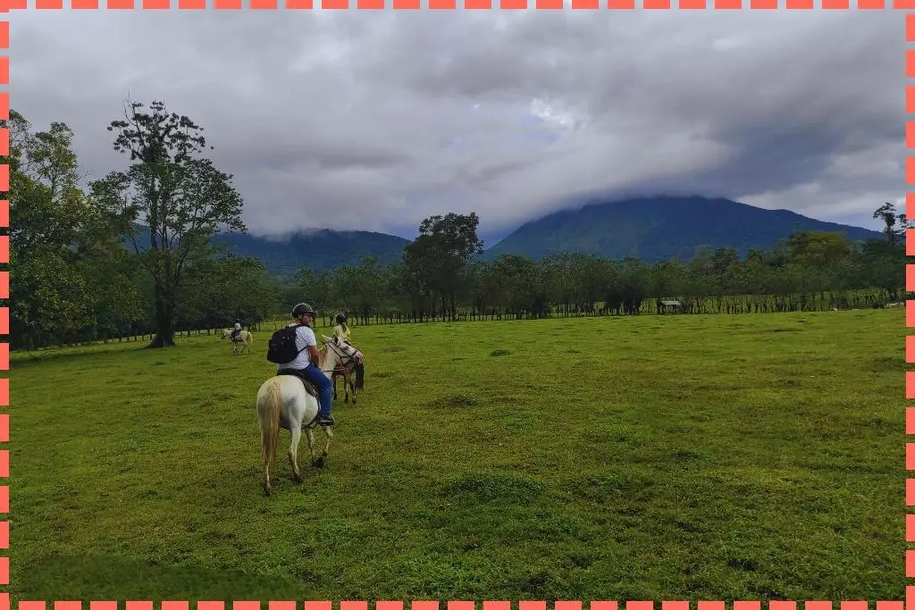 Familia Watson montando a caballo en el campo con el Volcán La Fortuna en el fondo, aunque no es claramente visible debido a las nubes.