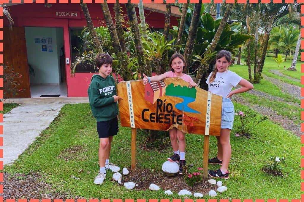 Ian, Sofi, y Vicky sonríen frente al cartel de entrada de la Posada Río Celeste, con la recepción y el hermoso jardín de fondo.