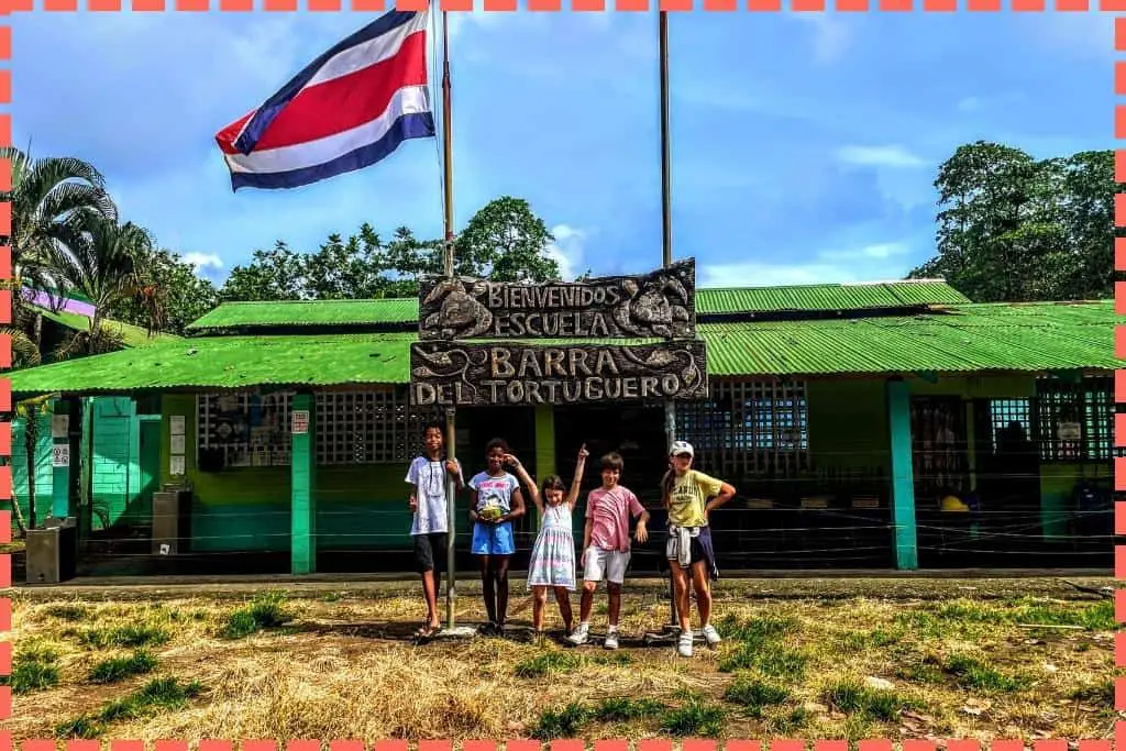 Niños visitantes y locales posando juntos con sonrisas amistosas en la entrada del colegio de Tortuguero, Costa Rica, destacando la unión cultural y el intercambio educativo.