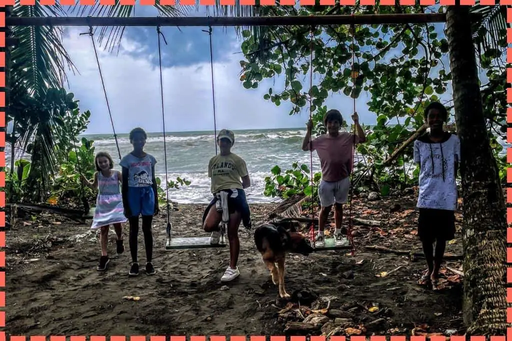Niños locales y visitantes disfrutando juntos de los columpios frente a la idílica playa de Tortuguero, con el sonido de las olas de fondo en un ambiente de alegría compartida.