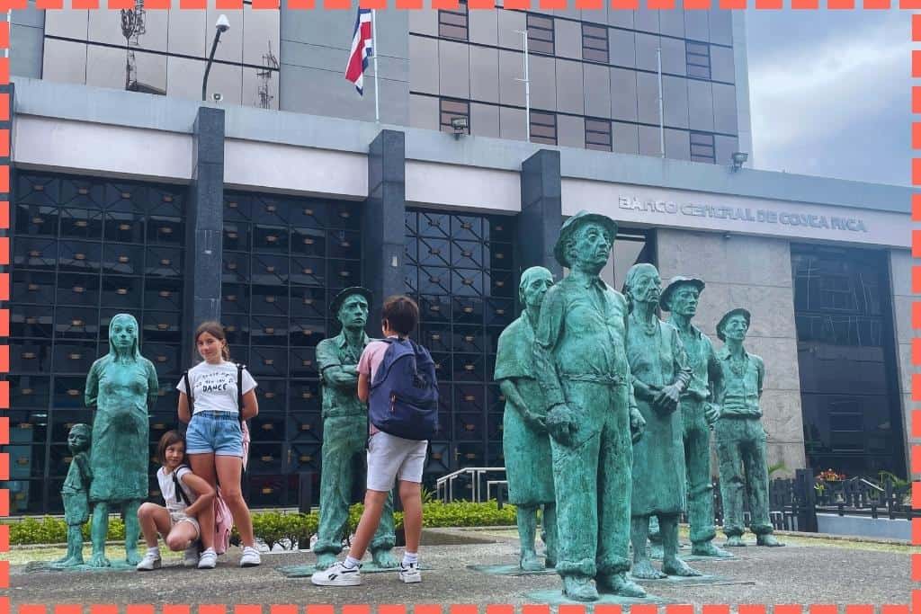 Tres hijos de Tere Watson interactuando con las estatuas de bronce 'Los Presentes' en San José, Costa Rica. Las estatuas, de tamaño similar al de los niños, se convierten en compañeros silenciosos en sus juegos imaginativos, creando una conexión entre el arte y la juventud.