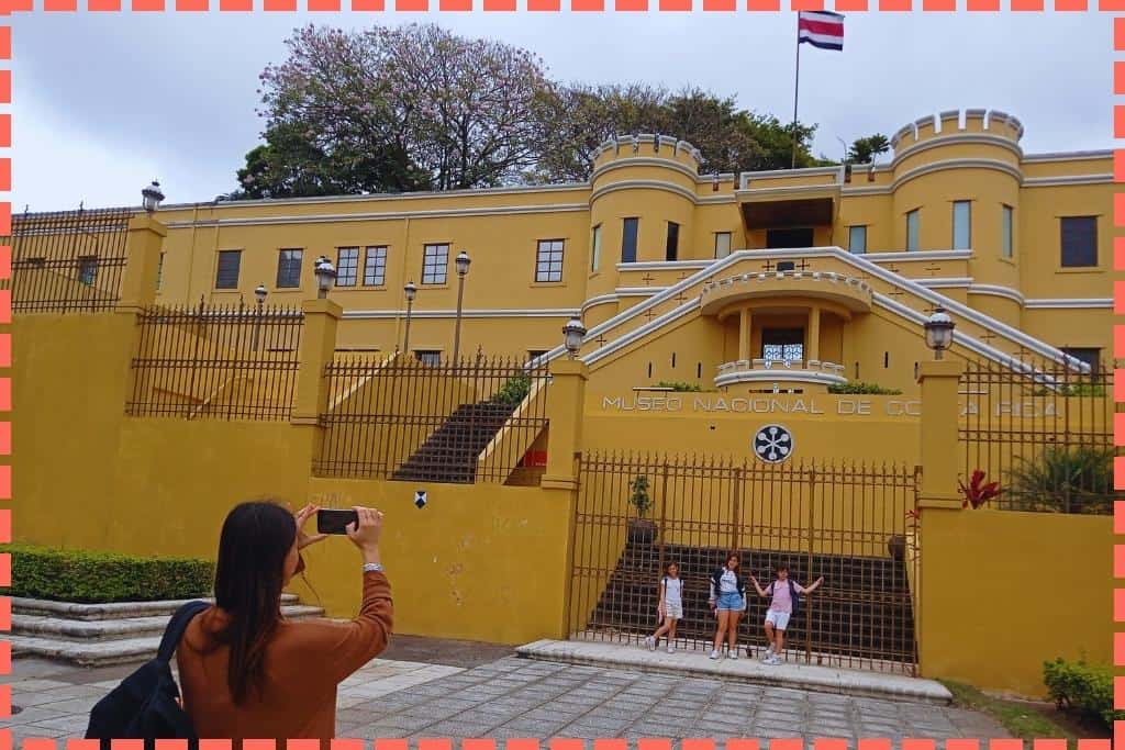 Tere Watson tomando una fotografía de sus tres hijos frente al Museo Nacional de Costa Rica. Los niños posan sonrientes en el exterior del museo, un lugar emblemático en San José que ofrece una inmersión en la rica historia y cultura del país