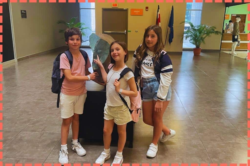 Tres niños, hijos de Tere Watson, en el hall de entrada del Museo del Jade en San José, Costa Rica, señalando un trozo de piedra de jade expuesta, de la que se han elaborado varios objetos en el museo, mostrando curiosidad y aprendizaje familiar en la ciudad.